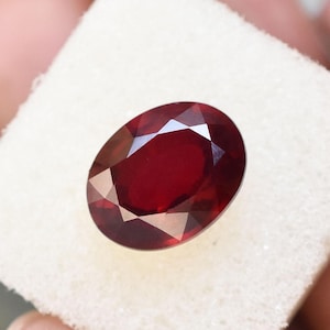 May include: A faceted, oval-shaped, deep red gemstone. The gem is set against a white background, showcasing its polished surfaces and internal reflections. The gemstone's rich color and cut create a brilliant display.