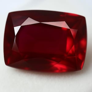 May include: A faceted, cushion-cut ruby gemstone. The gem is a deep, saturated red color with sharp, reflective facets. The shape is rectangular with rounded corners, and the stone is set against a white background.