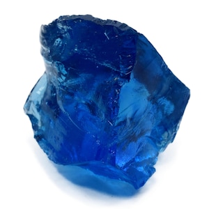 Op de afbeelding: Een levendig, onregelmatig gevormd, diepblauw kristal of edelsteen. Het doorschijnende materiaal heeft een glasachtige uitstraling, met facetten en een ruw, ongelijkmatig oppervlak. De edelsteen staat tegen een witte achtergrond, wat de kleur en textuur benadrukt.