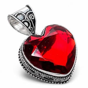 May include: A heart-shaped pendant featuring a large, faceted red gemstone set in a silver frame. The silver setting displays intricate scrollwork and beaded detailing along the edges, with a prominent, ornate bail at the top for attachment.