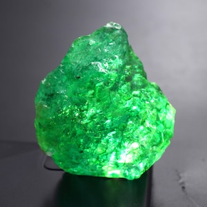 May include: A vibrant, raw emerald gemstone with a rough, textured surface. The gem is a deep, saturated green color, with areas of light and shadow that create a sparkling effect. The overall shape is irregular and natural, suggesting it is in its original, unpolished form.