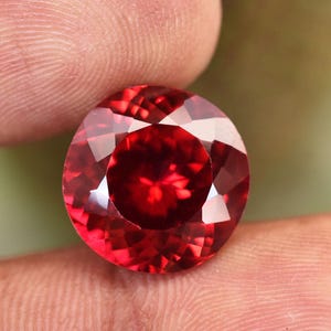 May include: A round, faceted, deep red gemstone. The gem is held between fingers, highlighting its brilliant cut and rich colour. The facets reflect light, creating a sparkling effect. The gemstone appears to be a ruby or similar precious stone.