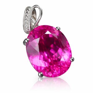 May include: A vibrant pink gemstone pendant with a silver bail. The oval-cut stone is held in place by four silver prongs. The bail is adorned with small, sparkling accents. The pendant is set against a white background.
