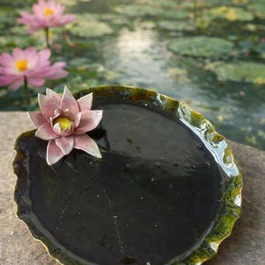 May include: A ceramic lily pad dish with a pink and yellow flower. The dish is dark green with a lighter green rim. The background shows a pond with lily pads and pink lotus flowers.