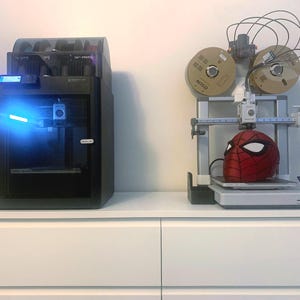 May include: Two 3D printers on a white surface. One is black with a blue light, the other is silver and printing a red and black Spider-Man mask. Two spools of filament are mounted above the mask.