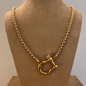 May include: A gold-colored necklace with a unique U-shaped clasp. The chain is made of small, rectangular links. The necklace is displayed on a tan mannequin. The clasp has a heart-shaped detail.