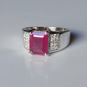 May include: A silver-toned ring featuring a large, rectangular, deep pink gemstone set in the center. The band is accented on both sides of the central stone with two vertical rows of small, clear, sparkling stones set into the metal surface.