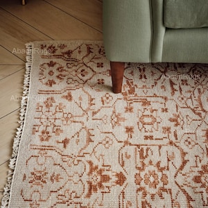 May include: Beige and rust-colored patterned rug with a distressed, vintage look. The rug features a detailed floral and geometric design, with a fringed edge. The rug is partially under a green armchair.