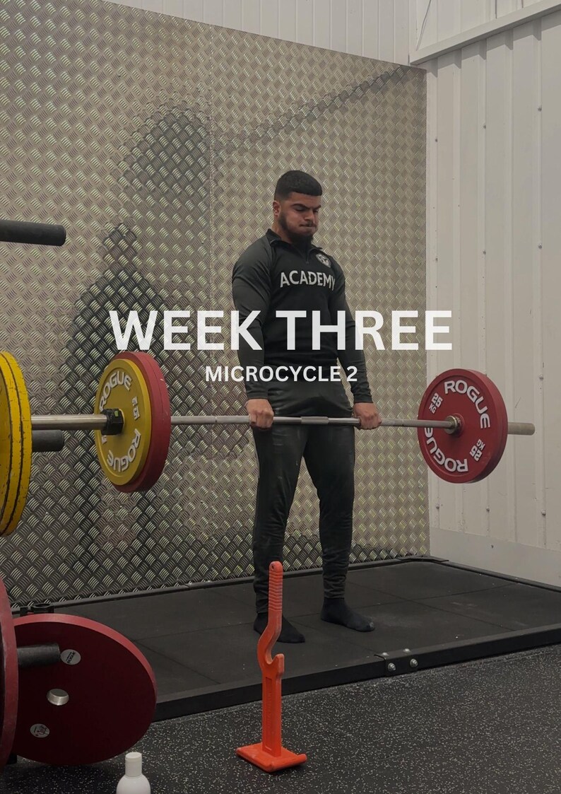 Puede incluir: Una persona realizando un peso muerto con una barra cargada con discos rojos y amarillos. El texto "WEEK THREE MICROCYCLE 2" se muestra en blanco. La persona lleva una camiseta negra de manga larga y pantalones negros.