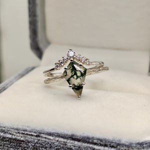 May include: A two-piece ring set featuring a silver band with a diamond-shaped moss agate stone and a delicate crown-shaped band with small clear stones. The moss agate has green and brown inclusions. The rings are displayed in a gray ring box.