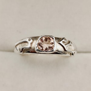 Puede incluir: Un anillo de plata con una piedra preciosa color melocotón en un engaste cuadrado. La banda tiene un diseño orgánico y texturizado con pequeñas piedras de acento transparentes. El diseño del anillo está inspirado en la naturaleza.