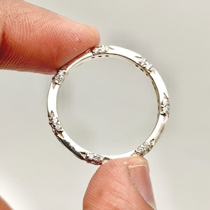 May include: A silver ring with a repeating pattern of small, clear gemstones. The ring is held between two fingers, showcasing its circular design and the sparkle of the stones. The ring's band has a textured, detailed design.