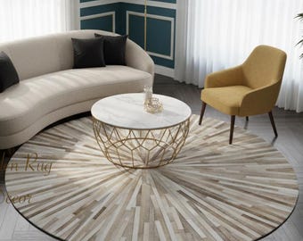 May include: A round area rug with a sunburst pattern in shades of beige, cream, and brown, placed in a living room with a curved beige sofa, black throw pillows, a mustard yellow armchair, and a round coffee table with a white marble top.