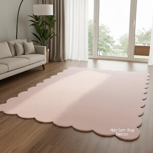 Puede incluir: Una alfombra grande, de color rosa claro, con un borde festoneado. La alfombra es rectangular con esquinas redondeadas y est&aacute; colocada sobre un suelo de madera. La alfombra est&aacute; en una habitaci&oacute;n con un sof&aacute;, una planta y una ventana grande.
