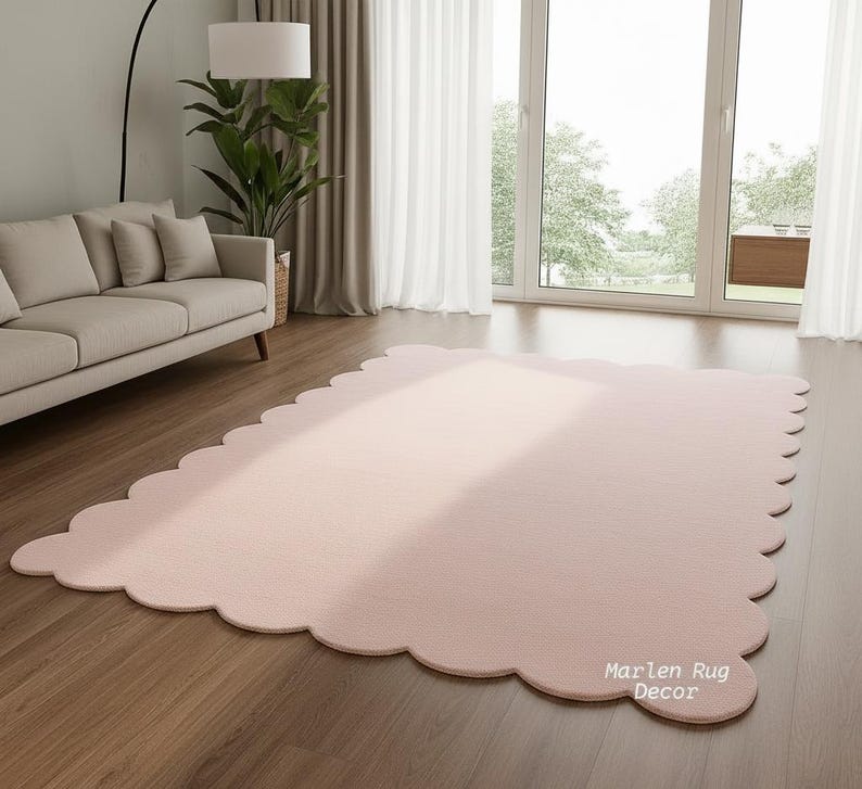 Puede incluir: Una alfombra grande, de color rosa claro, con un borde festoneado. La alfombra es rectangular con esquinas redondeadas y est&aacute; colocada sobre un suelo de madera. La alfombra est&aacute; en una habitaci&oacute;n con un sof&aacute;, una planta y una ventana grande.