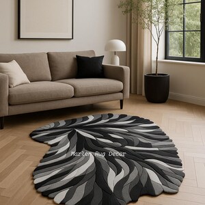 May include: A modern, abstract rug in shades of gray and black, with a swirling, organic pattern. The rug is placed on a light-colored wooden floor in a living room setting. The text "Marlen Rug Decor" is visible on the rug.