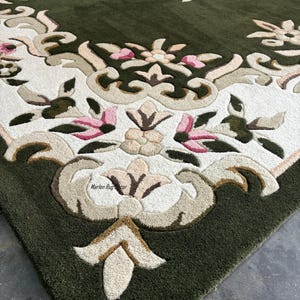 May include: Close-up of a green area rug with an ornate floral design. The rug features a central medallion and border with cream, tan, and pink floral patterns. The rug's texture is visible, with a dense, plush pile.
