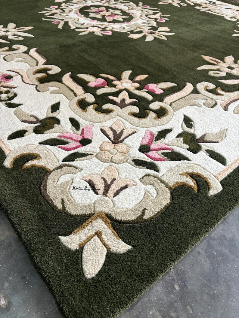 May include: Close-up of a green area rug with an ornate floral design. The rug features a central medallion and border with cream, tan, and pink floral patterns. The rug's texture is visible, with a dense, plush pile.