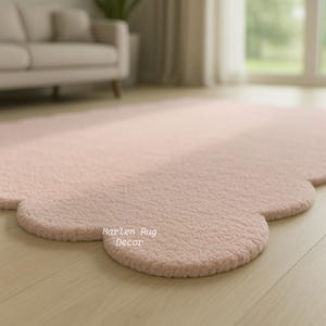 Puede incluir: Una alfombra rosa claro en forma de nube con una superficie texturizada. La alfombra est&aacute; sobre un suelo de madera clara. Las palabras "Marlen Rug Decor" est&aacute;n impresas en la alfombra.