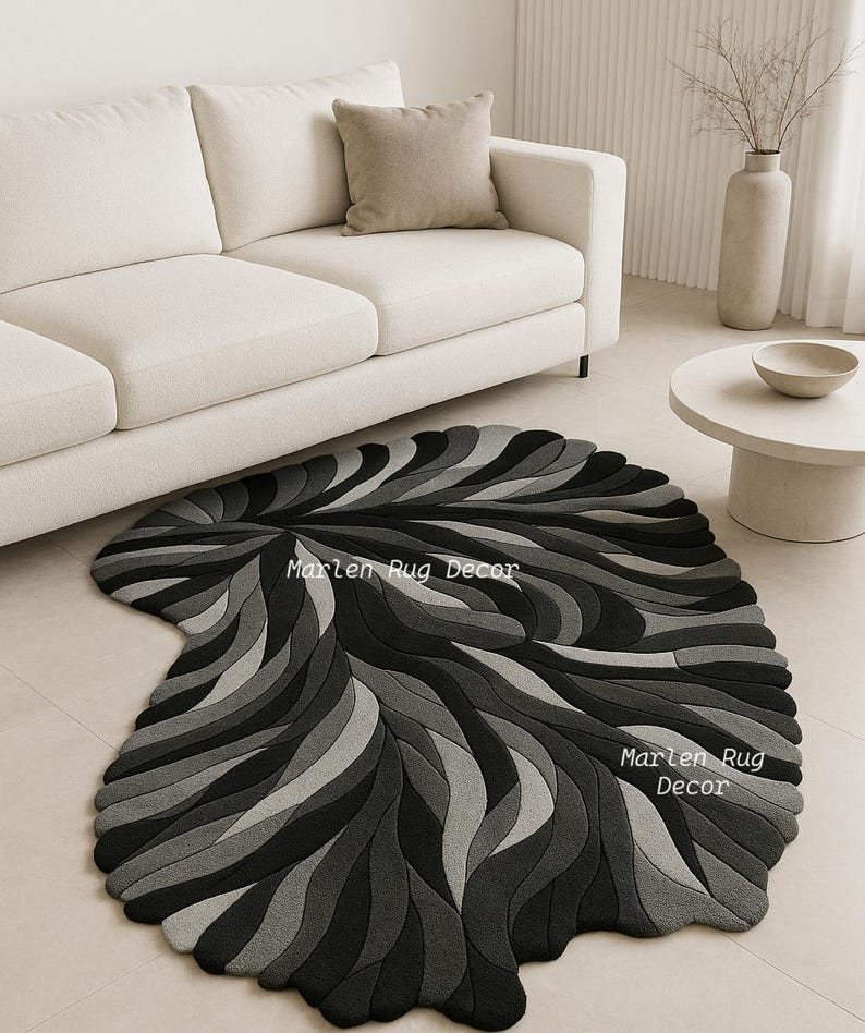 May include: A modern, abstract rug in shades of gray and black, featuring a leaf-like design. The rug is placed on a light-colored tile floor, with a white sofa and neutral-toned decor in the background. The text "Marlen Rug Decor" is visible on the rug.