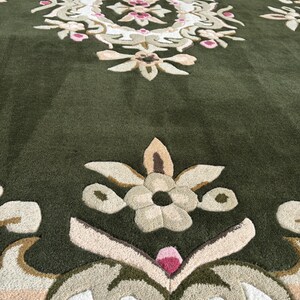 May include: Close-up of a green rug with an intricate floral design. The rug features a central medallion with pink and white floral elements, surrounded by beige and green patterns. The text "Marlen Rug Decor" is visible.