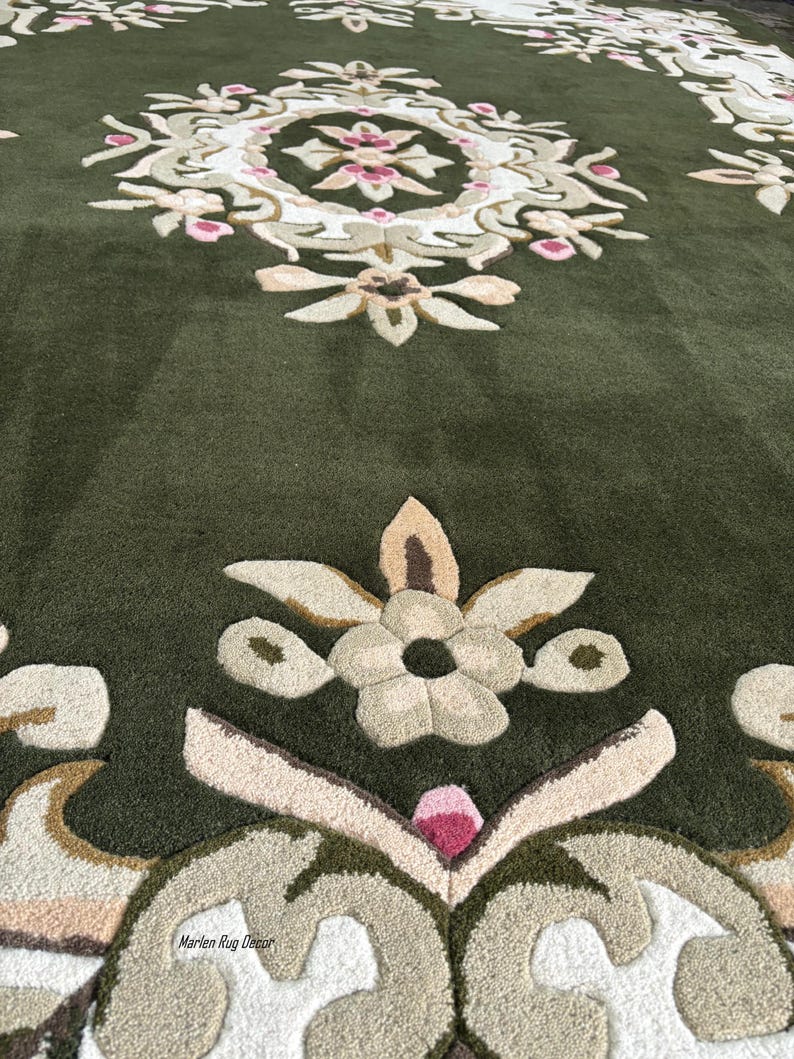 May include: Close-up of a green rug with an intricate floral design. The rug features a central medallion with pink and white floral elements, surrounded by beige and green patterns. The text "Marlen Rug Decor" is visible.