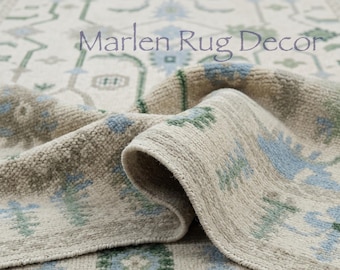 May include: A woven area rug featuring a cream-colored base with a repeating pattern of light blue and sage green floral motifs. The text Marlen Rug Decor is printed in a dark purple font across the center of the folded, textured fabric surface.