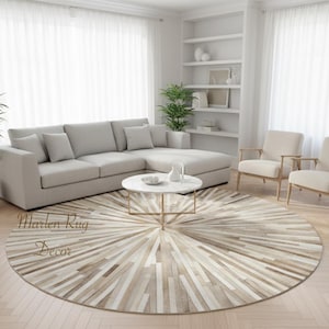 Op de afbeelding: Een ronde vloerkleed met een sunburst-ontwerp in beige, crème en bruine tinten. Het vloerkleed ligt in een woonkamer met een lichtgrijze hoekbank, twee fauteuils en een salontafel met marmeren blad. De kamer heeft witte gordijnen en een boekenkast.