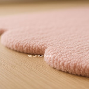 Puede incluir: Primer plano de una alfombra rosa suave con una forma redondeada &uacute;nica. La alfombra tiene una superficie texturizada y afelpada y est&aacute; sobre un fondo de madera clara. El texto "Marlen Rug Decor" es visible.