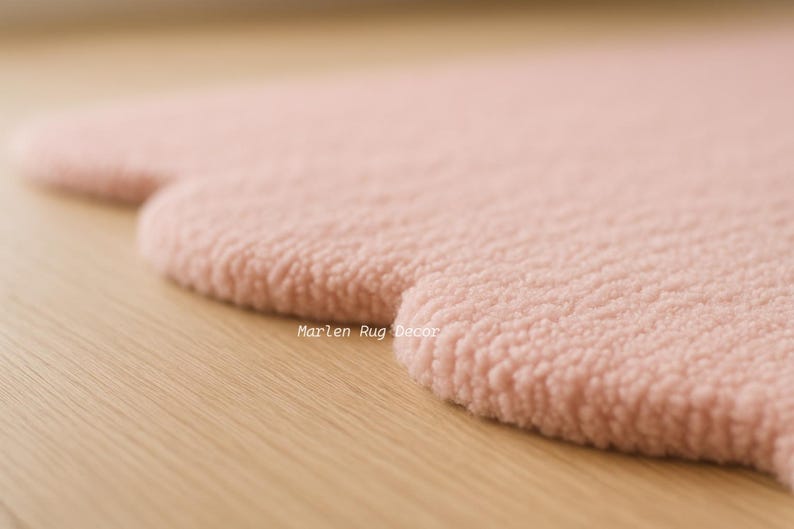 Puede incluir: Primer plano de una alfombra rosa suave con una forma redondeada &uacute;nica. La alfombra tiene una superficie texturizada y afelpada y est&aacute; sobre un fondo de madera clara. El texto "Marlen Rug Decor" es visible.