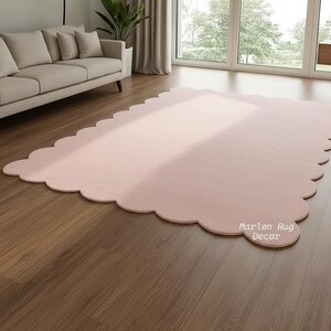 Puede incluir: Una alfombra grande, de color rosa claro, con borde festoneado se muestra sobre un suelo de madera. La alfombra est&aacute; en un sal&oacute;n, con un sof&aacute; y una ventana visibles en el fondo. El texto "Marlen Rug Decor" est&aacute; impreso en la alfombra.