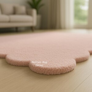 Puede incluir: Una alfombra rosa claro con forma de nube y superficie texturizada. La alfombra est&aacute; sobre un suelo de madera clara. El texto "Marlen Rug Decor" es visible en la alfombra.