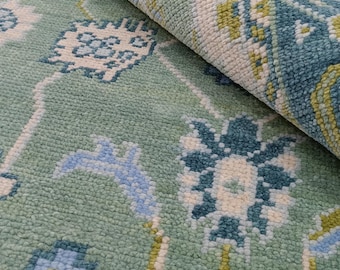 Könnte beinhalten: Nahaufnahme eines aufgerollten Teppichs mit floralem Muster. Der Teppich hat einen hellgrünen Hintergrund mit blauen, weißen und gelben floralen Designs. Die Textur des Teppichs ist sichtbar, und der aufgerollte Teil zeigt die Unterseite des Teppichs.
