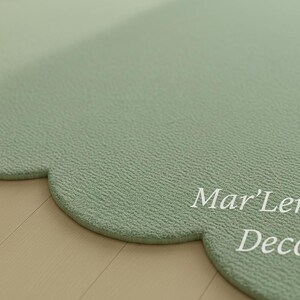 Puede incluir: Una alfombra verde salvia en forma de nube con una superficie texturizada. La alfombra tiene un borde festoneado y las palabras "Mar'Len Deco" est&aacute;n impresas en blanco en la superficie. La alfombra est&aacute; colocada sobre un suelo de madera clara.