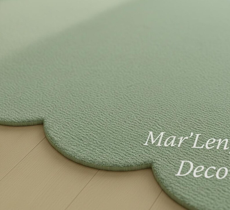 Puede incluir: Una alfombra verde salvia en forma de nube con una superficie texturizada. La alfombra tiene un borde festoneado y las palabras "Mar'Len Deco" est&aacute;n impresas en blanco en la superficie. La alfombra est&aacute; colocada sobre un suelo de madera clara.