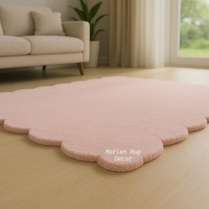 Puede incluir: Una alfombra rosa claro con bordes festoneados sobre un suelo de madera. La alfombra tiene una textura suave y afelpada y est&aacute; en un sal&oacute;n con un sof&aacute; beige y luz natural.