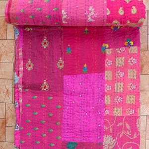 May include: A rolled-up, patchwork quilt in shades of pink and fuchsia. The quilt features embroidered floral and geometric patterns, with a variety of textures and stitching details. The quilt is made of multiple fabric squares.