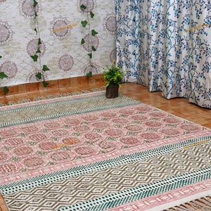 May include: A rectangular, patterned rug with a pink and green floral design. The rug has a geometric border and fringe on the ends. The rug is placed on a tiled floor, with floral curtains and a decorative wall in the background.