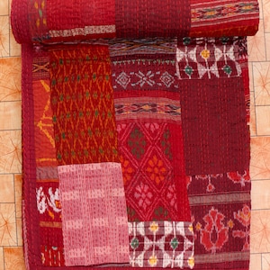 Puede incluir: Un edredón de retazos en tonos de rojo, burdeos y granate. El edredón está hecho de varios patrones de tela, incluyendo diseños geométricos y florales. El edredón está enrollado en la parte superior, mostrando las diferentes texturas y colores.