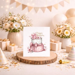 May include: A birthday greeting card featuring a two-tiered cake design with pink roses, a bow, and gold accents. The card has the words "Happy Birthday" in gold script. The card is on a wooden slice, surrounded by gifts and flowers.