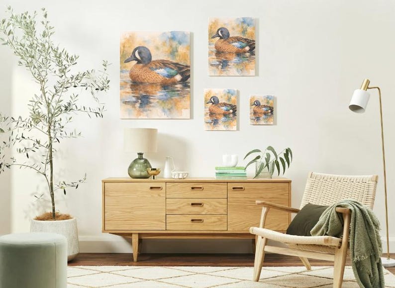 May include: A set of four canvas art prints featuring watercolor paintings of a duck on water. The duck has a blue head, brown body, and is set against a backdrop of muted blues and yellows. The prints vary in size and are displayed on a white wall.