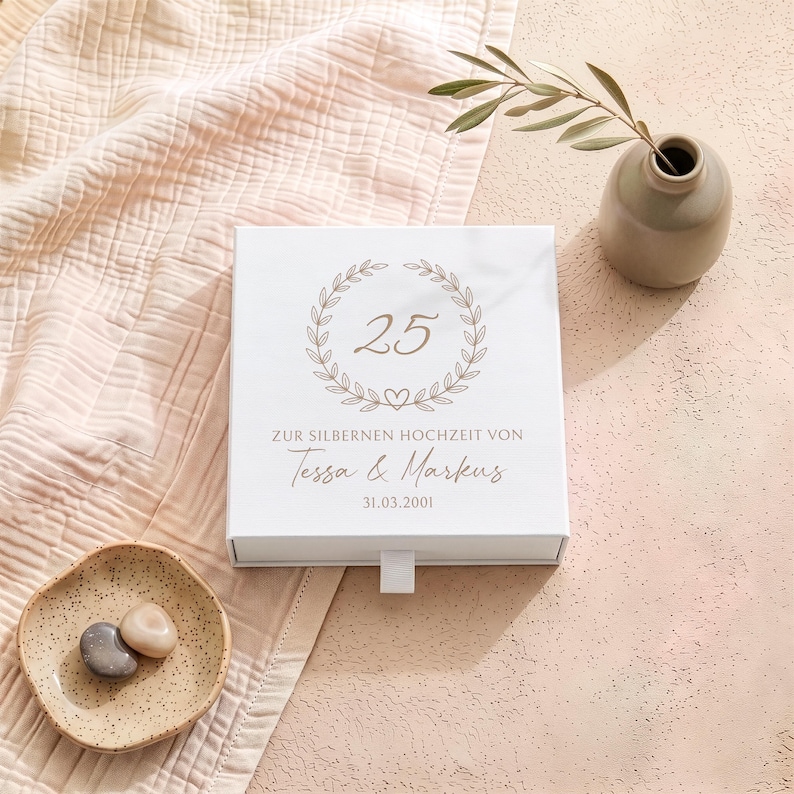 Pu&ograve; includere: Una scatola regalo quadrata bianca con una grafica a corona d'alloro color oro che circonda il numero 25, con il testo "ZUR SILBERNEN HOCHZEIT VON Tessa & Markus" e la data "31.03.2001" stampati in un carattere dorato corsivo e serif.