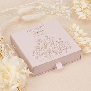 May include: A pale pink square box featuring a floral design and the name "Sophia" printed on the lid. The text "ZUR TAUFE VON" and the date "03.05.2026" are also printed on the lid. A pale pink ribbon is attached to the front of the box.