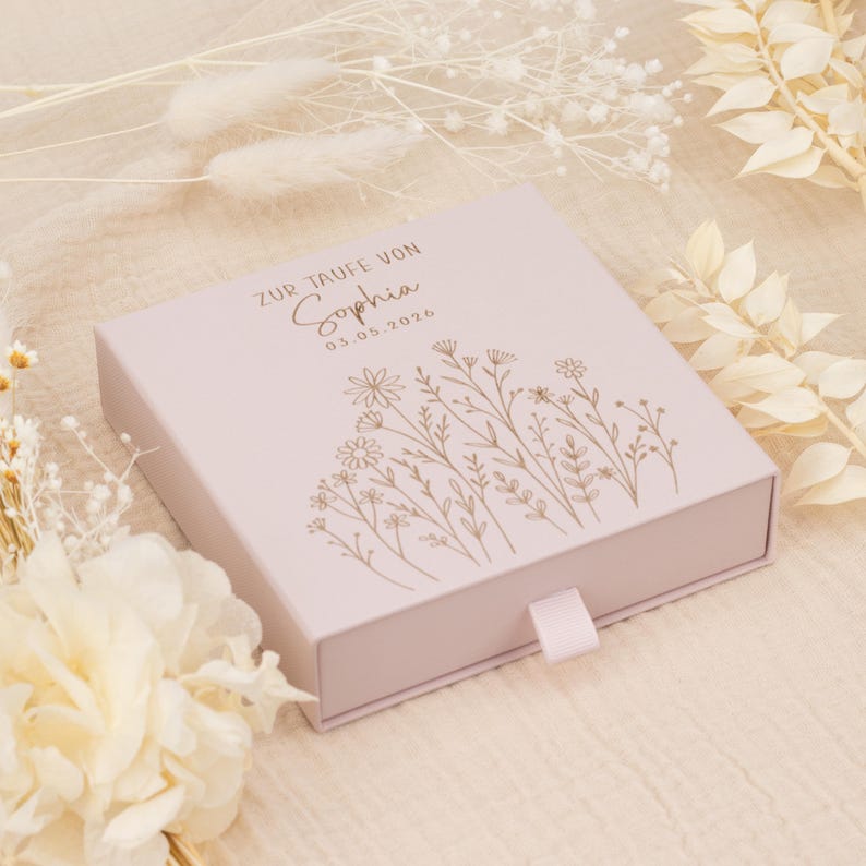 May include: A pale pink square box featuring a floral design and the name "Sophia" printed on the lid. The text "ZUR TAUFE VON" and the date "03.05.2026" are also printed on the lid. A pale pink ribbon is attached to the front of the box.