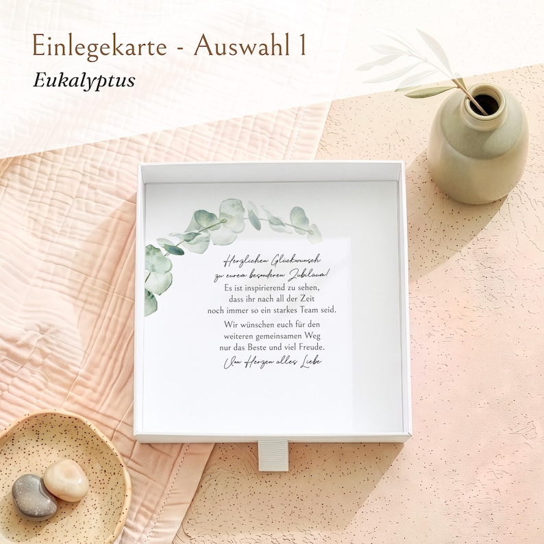 Pu&ograve; includere: Una scatola regalo quadrata bianca contenente un biglietto con un'illustrazione ad acquerello di un ramo di eucalipto e il testo in tedesco: Herzlichen Gl&uuml;ckwunsch zu eurem besonderen Jubil&auml;um! Es ist inspirierend zu sehen, dass ihr nach all der Zeit noch immer so ein starkes Team seid. Wir w&uuml;nschen euch f&uuml;r den weiteren gemeinsamen Weg nur das Beste und viel Freude. Im Herzen alles Liebe.