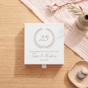 Pu&ograve; includere: Una scatola quadrata bianca con cassetto estraibile, decorata con una corona d'alloro color oro che circonda il numero 25, con il testo ZUR SILBERNEN HOCHZEIT VON Tessa & Markus e la data 31.03.2001 stampati in un carattere grigio scuro.