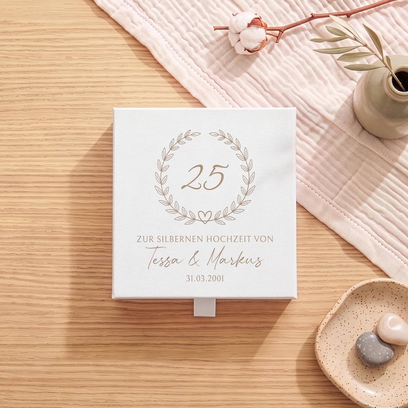Pu&ograve; includere: Una scatola quadrata bianca con cassetto estraibile, decorata con una corona d'alloro color oro che circonda il numero 25, con il testo ZUR SILBERNEN HOCHZEIT VON Tessa & Markus e la data 31.03.2001 stampati in un carattere grigio scuro.
