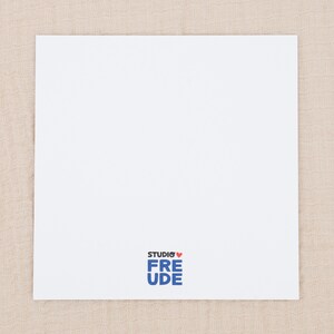 May include: A square white card with a smooth surface. At the bottom center, there is a logo featuring the text STUDIO in black, a small red heart icon, and the word FREUDE in bold blue letters stacked vertically below the heart.