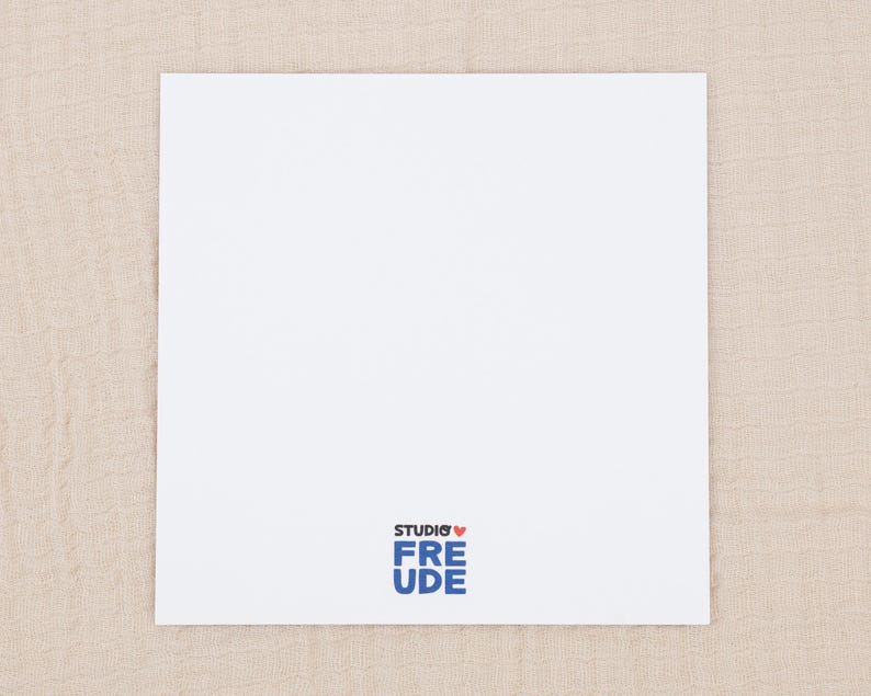 May include: A square white card with a smooth surface. At the bottom center, there is a logo featuring the text STUDIO in black, a small red heart icon, and the word FREUDE in bold blue letters stacked vertically below the heart.