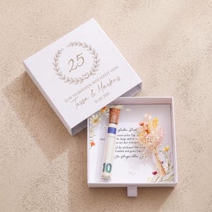 Pu&ograve; includere: Una scatola regalo quadrata bianca con coperchio scorrevole decorata con una corona d'alloro dorata, il numero 25 e il testo "ZUR SILBERNEN HOCHZEIT VON Tessa & Markus 31.03.2001". All'interno, una fiala di vetro con tappo in sughero e banconota.